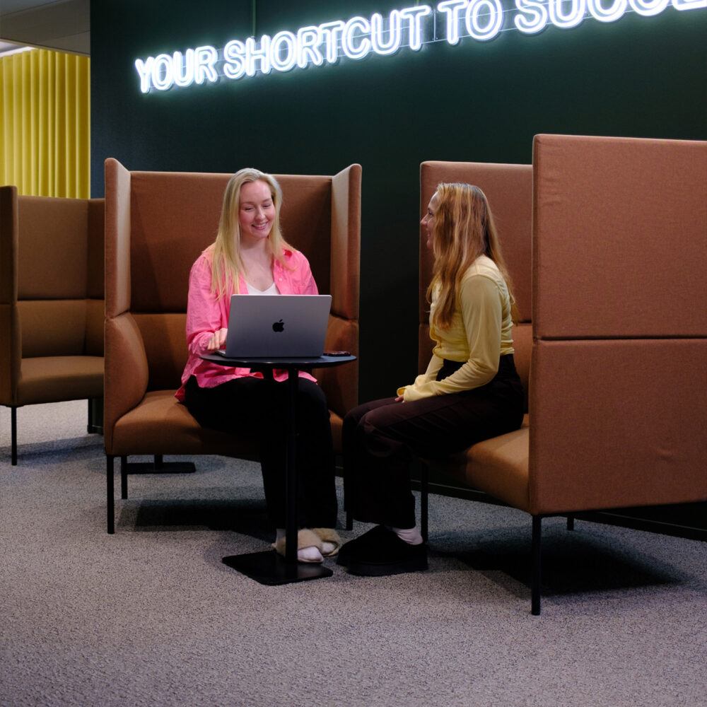 Kaksi henkilöä keskustelee kannettavan tietokoneen ääressä toimiston lounge-tilassa